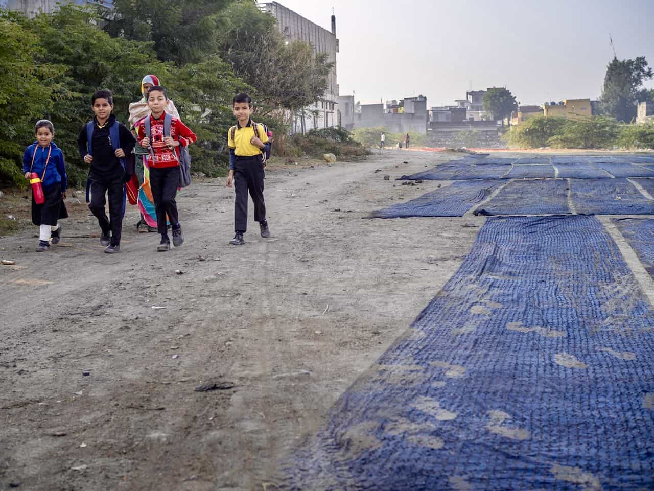 Group of schoolchildren walking along a dusty village road in India, accompanied by a woman in traditional attire, with blue-dyed fabric laid out on the ground to dry.
