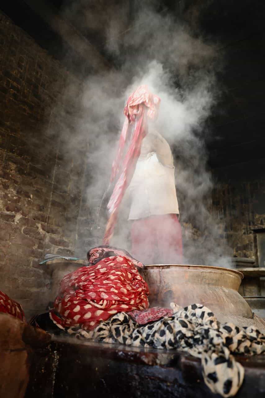 Man handling steaming, freshly dyed fabric from a large boiling vat inside a traditional textile workshop with exposed brick walls.