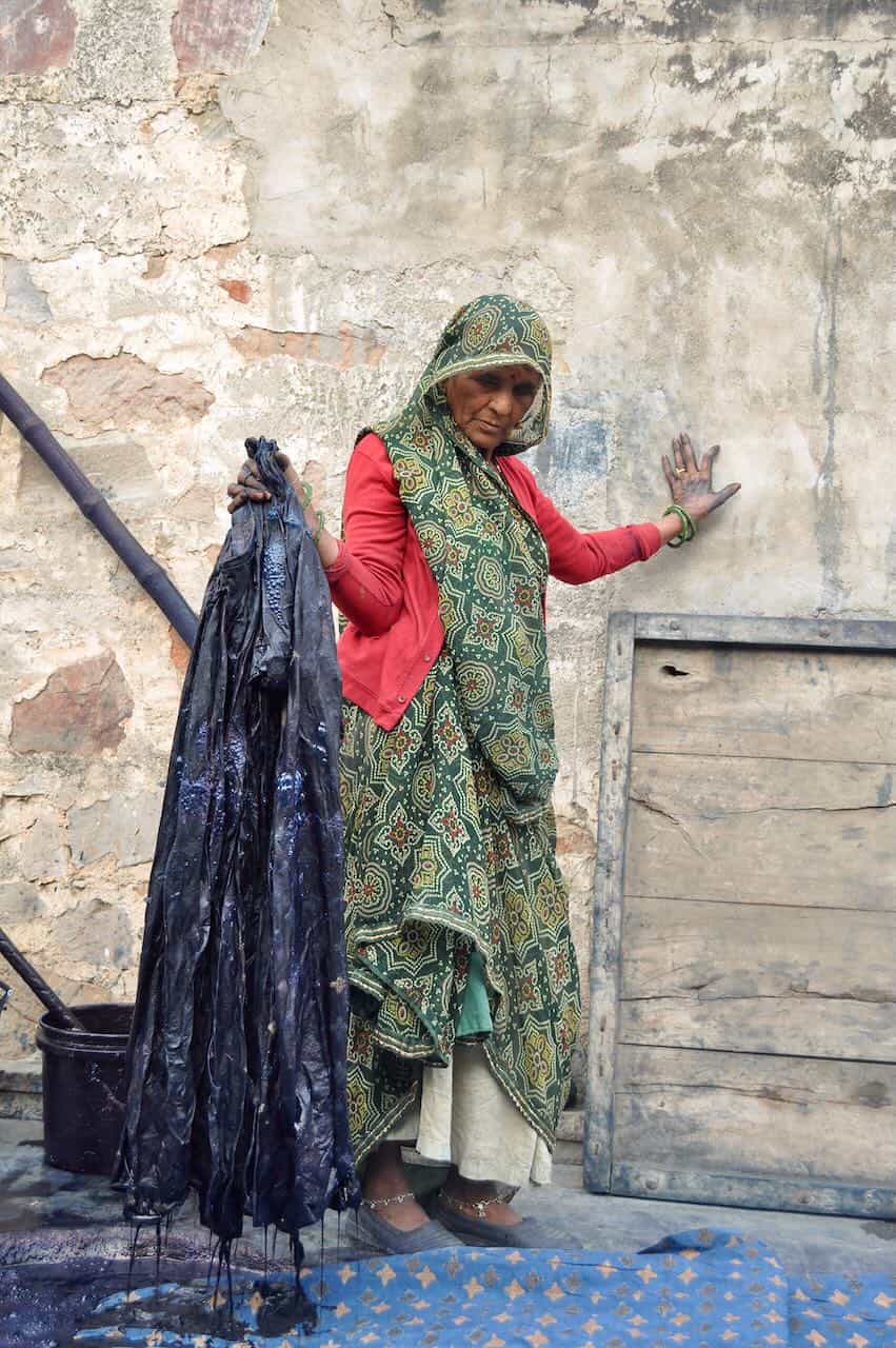 Elderly woman in colorful traditional clothing holding freshly dyed dark indigo fabric against a rustic wall in a hand-printing workshop.