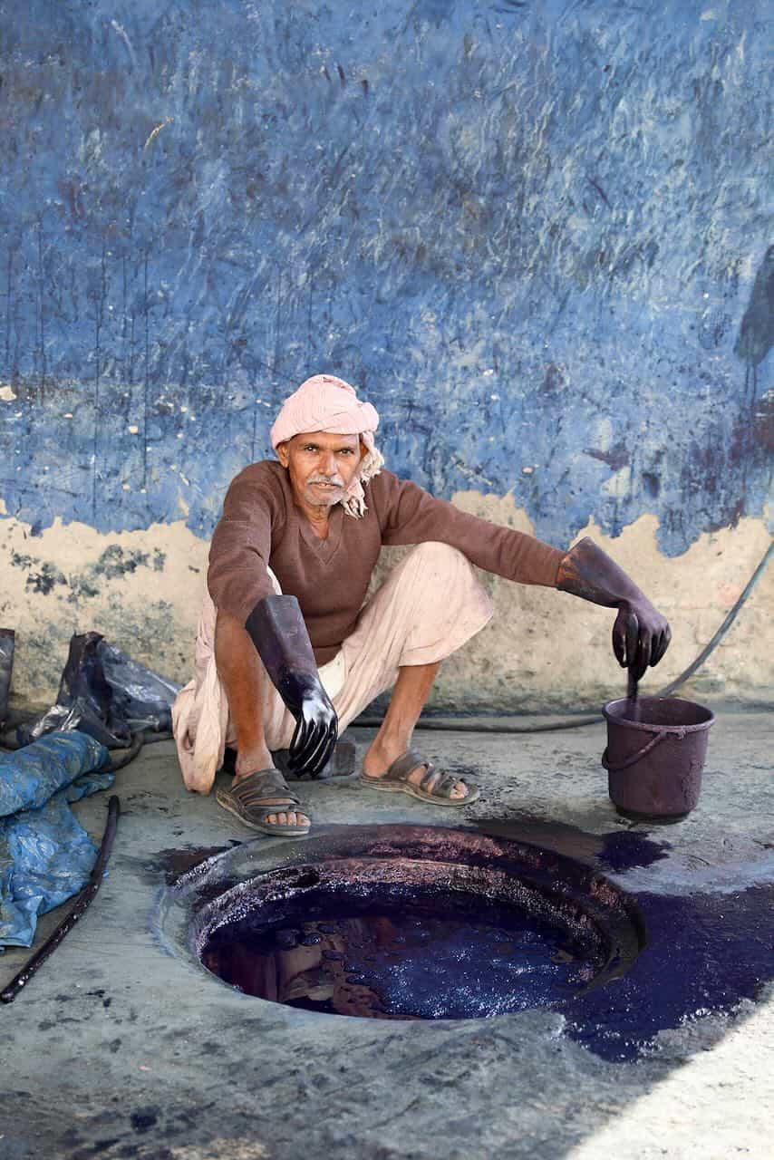 Man squatting beside an open indigo dye pit, wearing protective gloves and stirring fabric dye in a bucket, with splattered dye stains on the floor and wall.