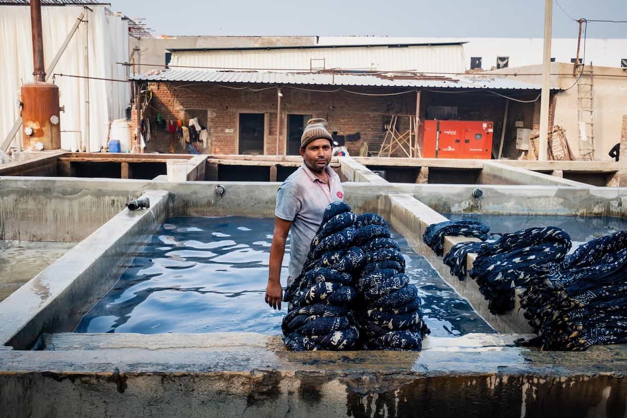 Man standing in a water-filled dyeing tank holding bundled textiles, surrounded by multiple concrete vats in an open-air textile processing area.