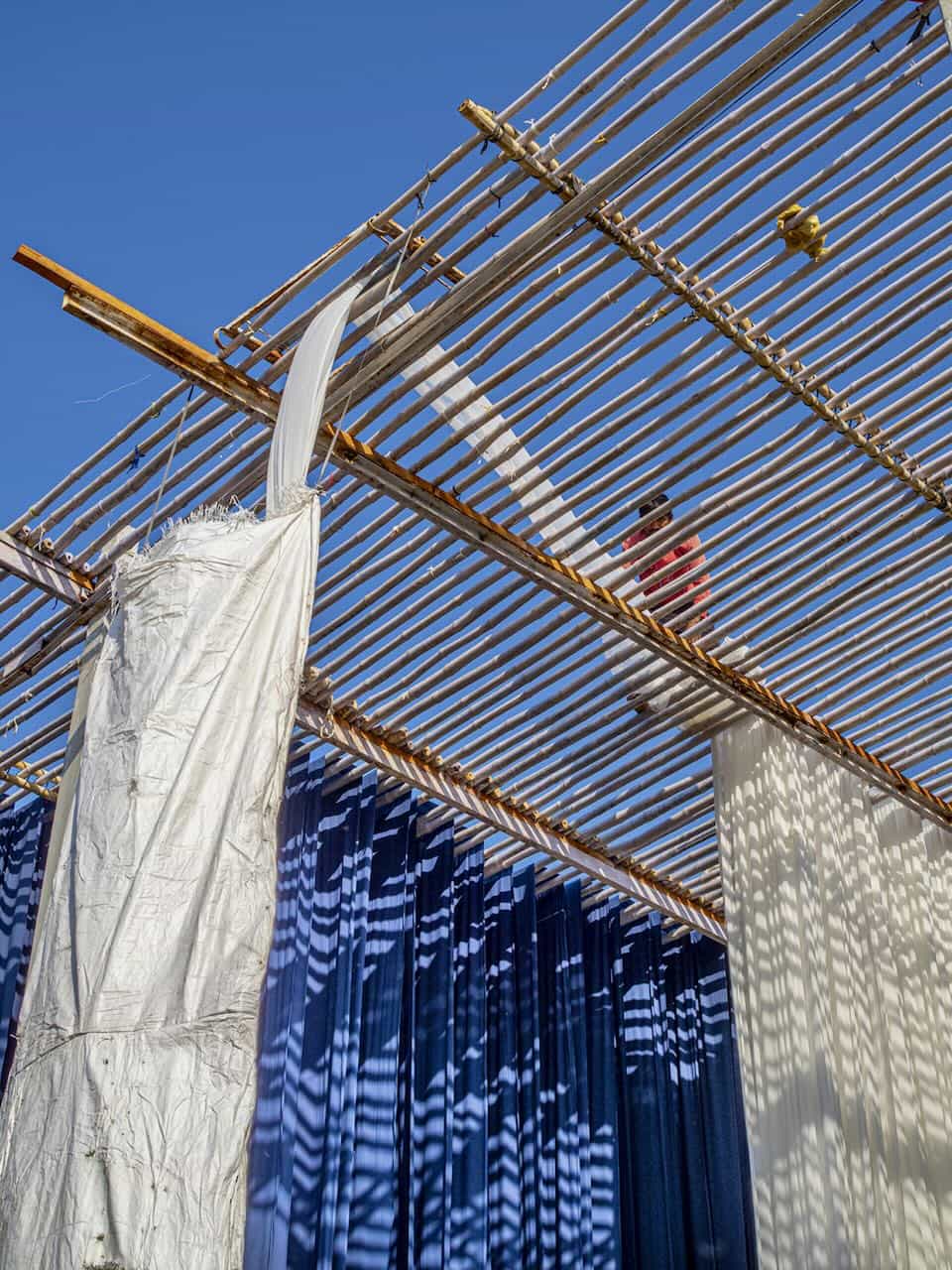 White and indigo-dyed fabrics hanging to dry under a bamboo structure in open sunlight, with a worker adjusting cloth above.