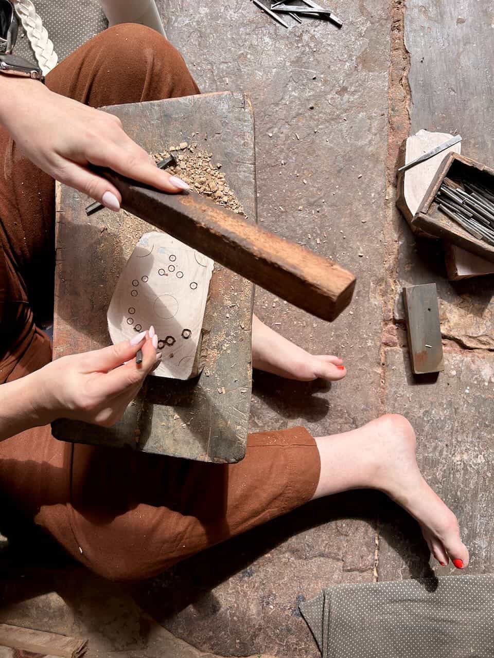 Close-up of a person carving circular patterns on a small wooden block while sitting on the floor.