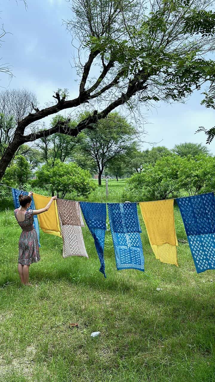 A woman hangs colorful naturally dyed fabrics—featuring bold patterns in yellow, indigo, and brown—on a clothesline under a large tree in a lush green outdoor setting.