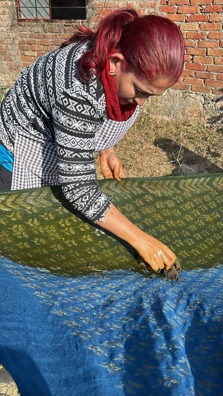 A workshop participant presses intricate block print patterns onto fabric using a traditional hand-carved wooden block, applying resist paste on a vibrant blue and green textile.
