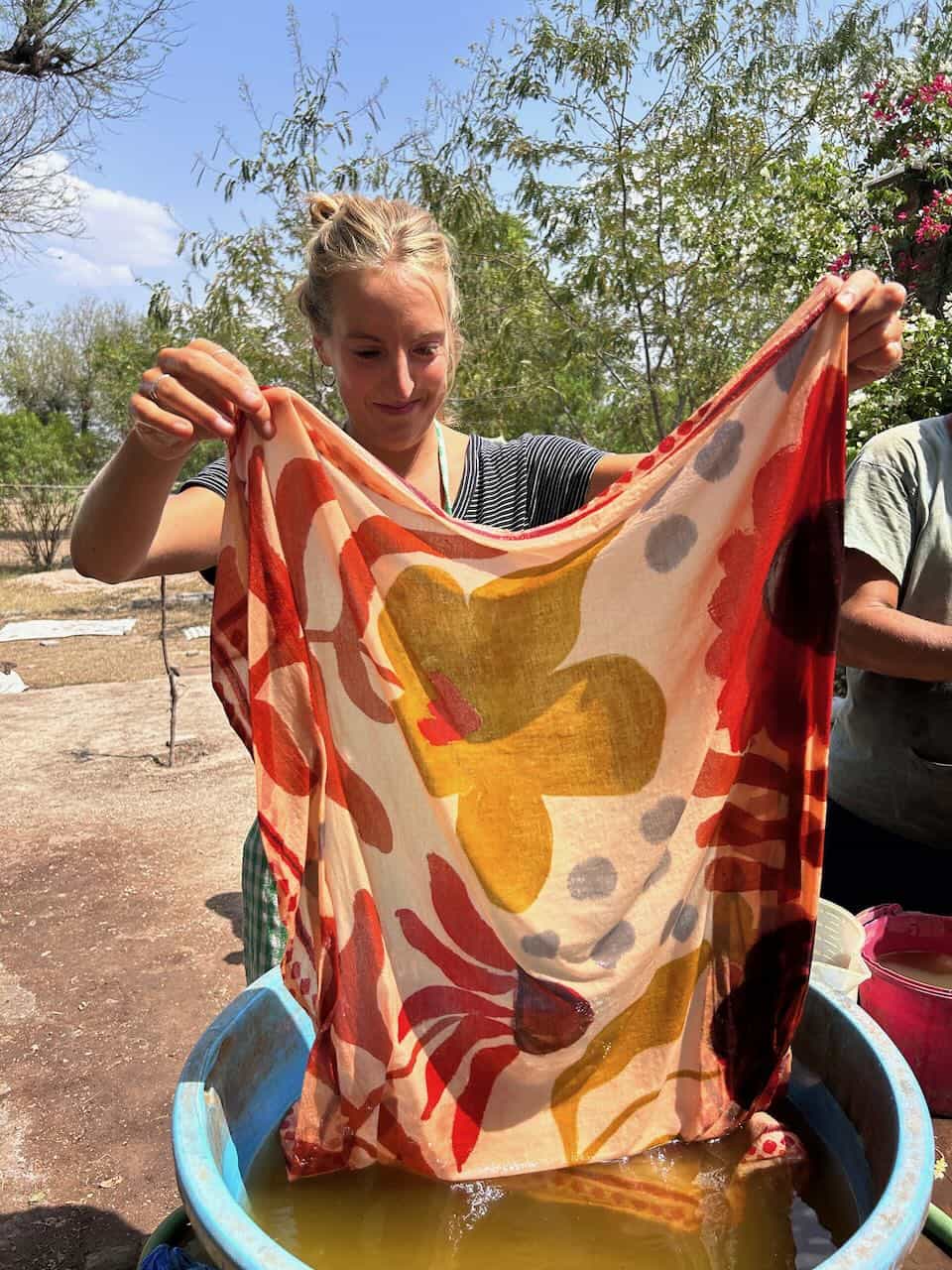 A smiling woman holds up a silk scarf dyed with natural pigments, revealing a vivid floral pattern in yellow, red, and orange hues after removing it from a dye bath.