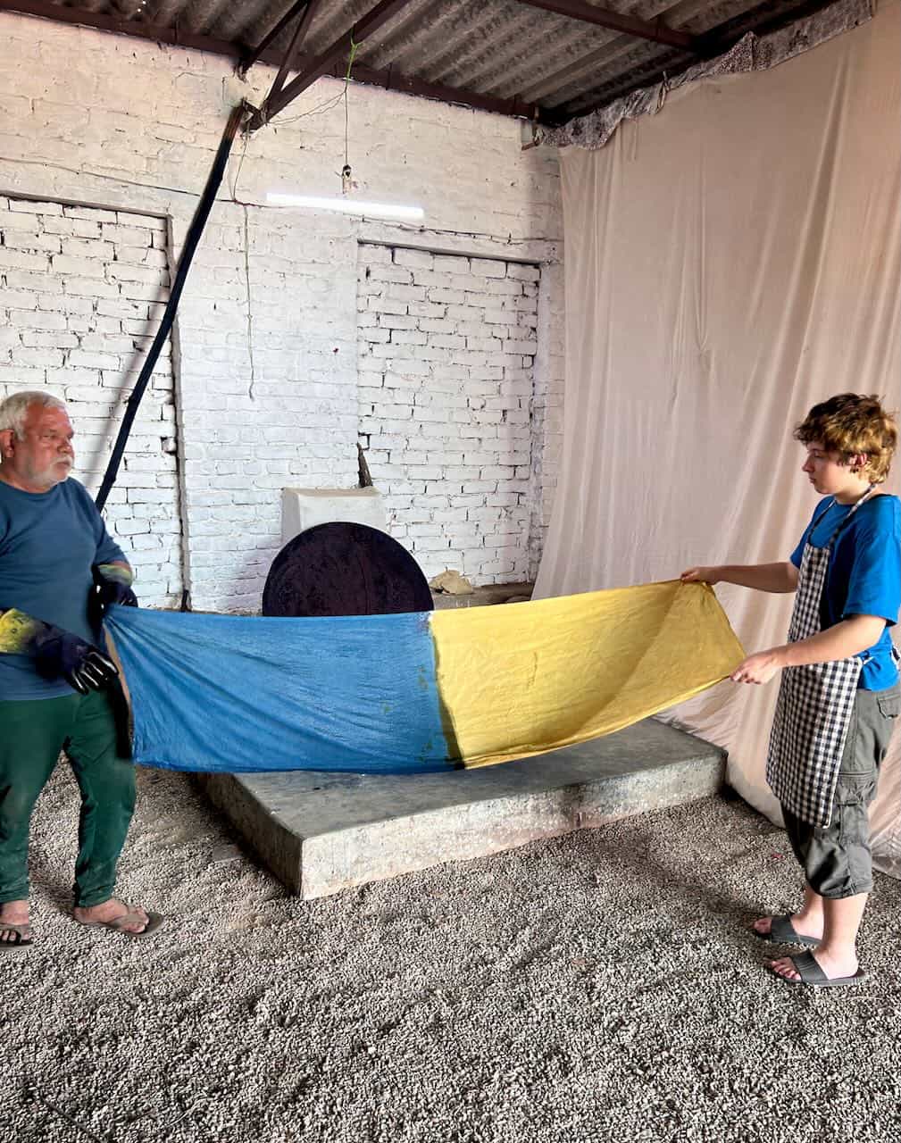 Two participants stretch out a large piece of fabric dyed in blue and yellow halves, displaying a color-blocking technique inside a rustic indoor dyeing workshop.
