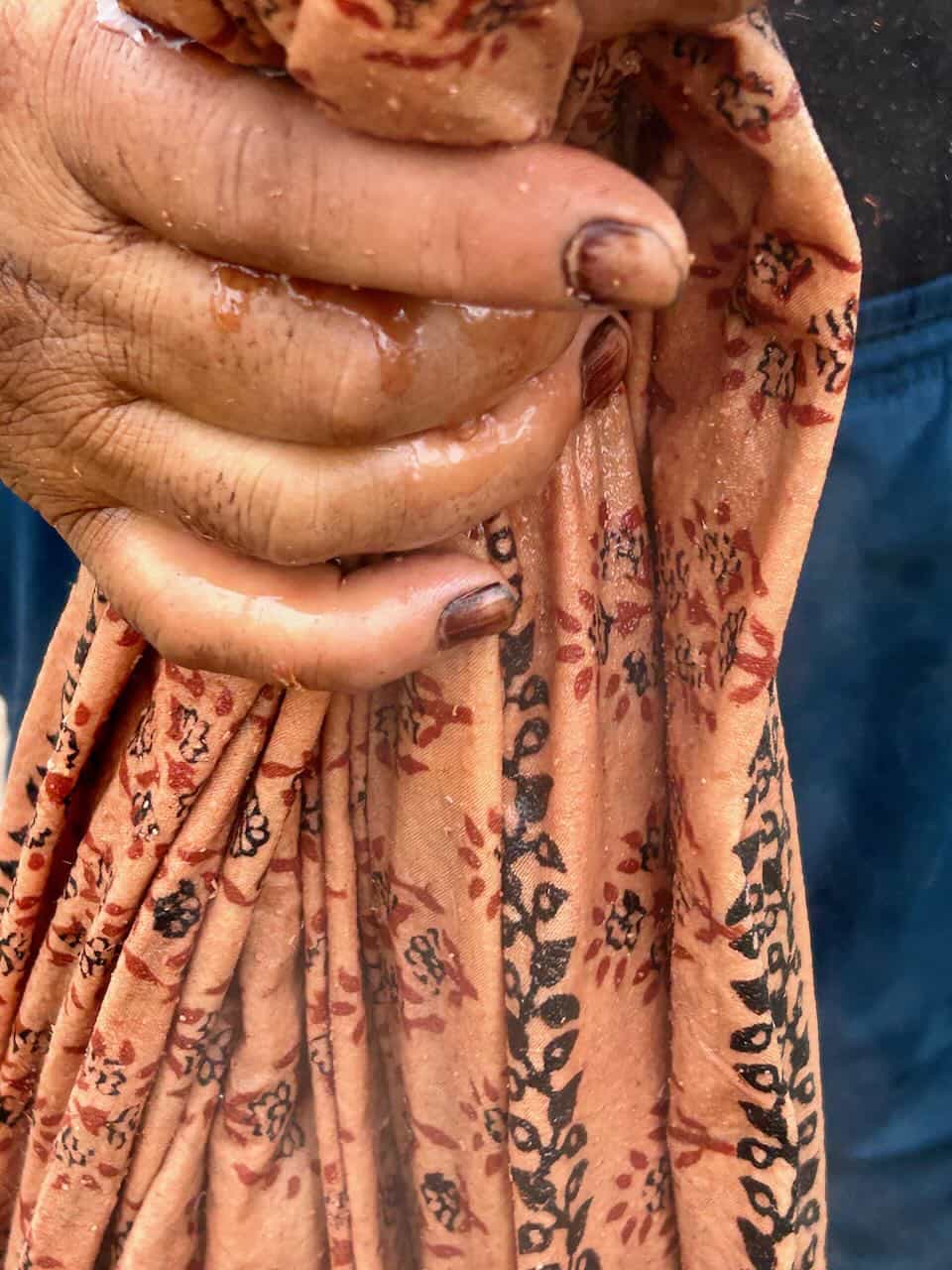 Close-up of a person's hand squeezing wet, naturally dyed fabric with intricate floral block print patterns in red and black on an earthy peach background.