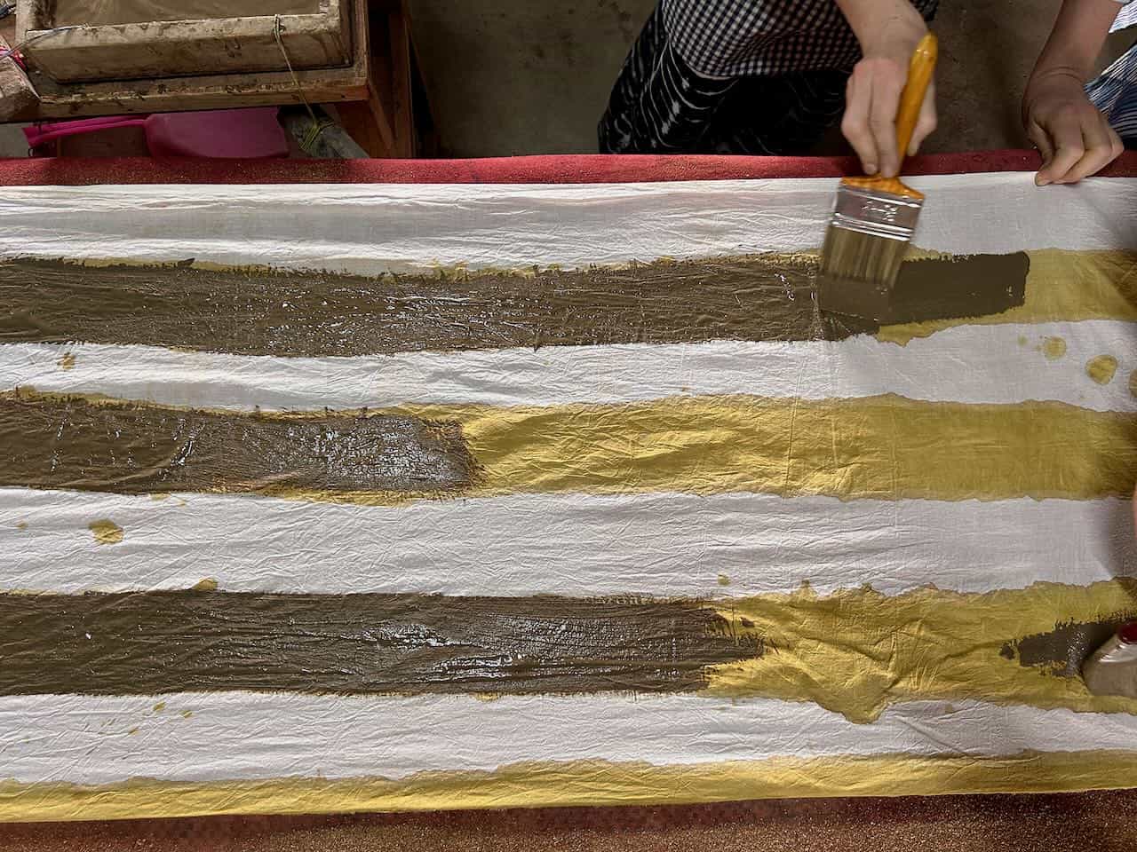 Person using a wide brush to apply natural dye in dark and golden yellow stripes onto white fabric during a traditional textile workshop.
