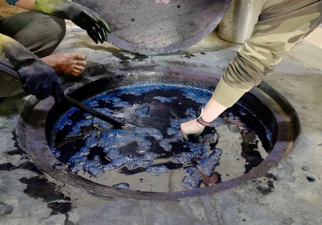 Hands stirring a large indigo dye vat with protective gloves, as fabric is dipped into the deep blue dye bath during the natural dyeing process.