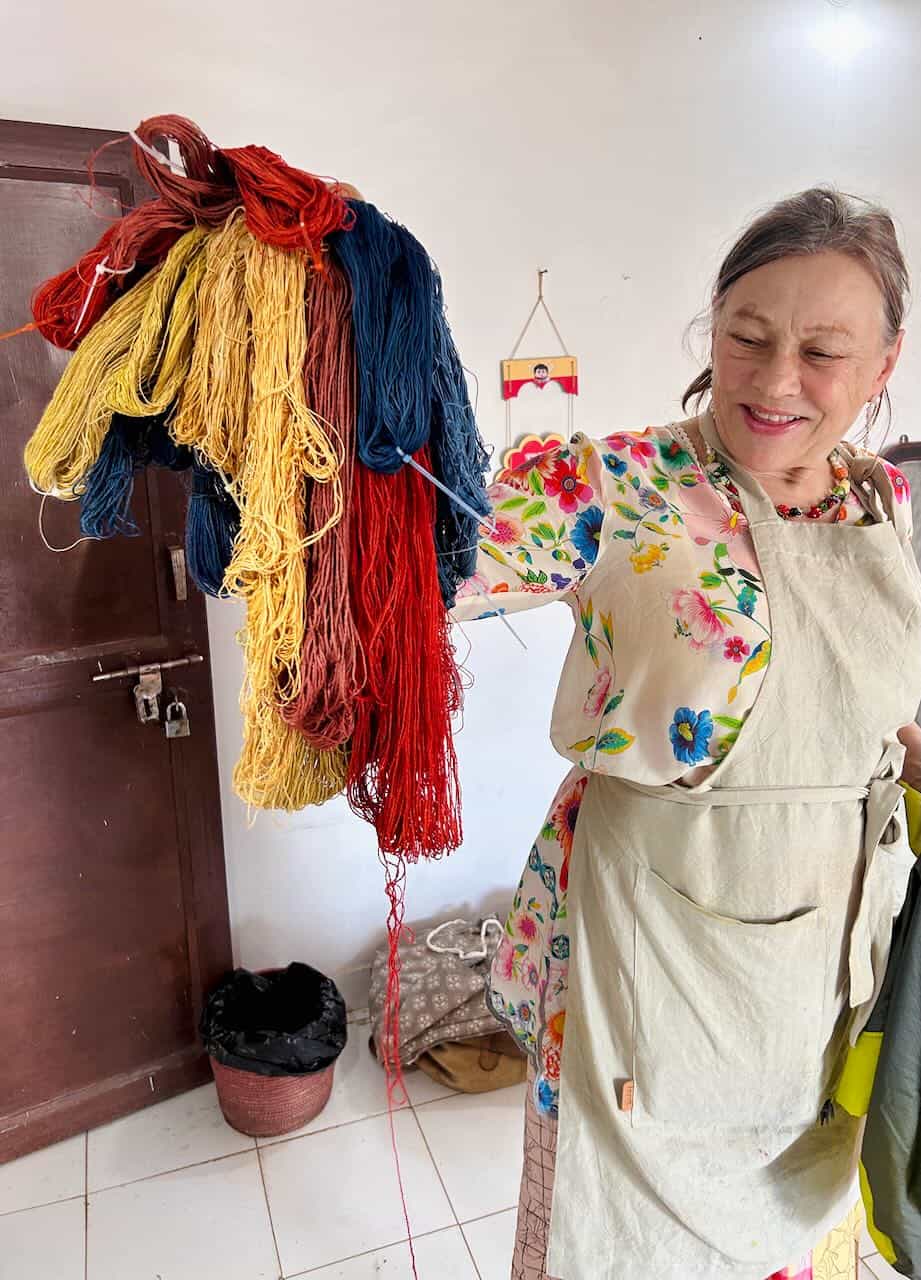 Smiling woman holding up vibrant bundles of hand-dyed yarn in red, yellow, blue, and brown shades inside a textile workshop space.