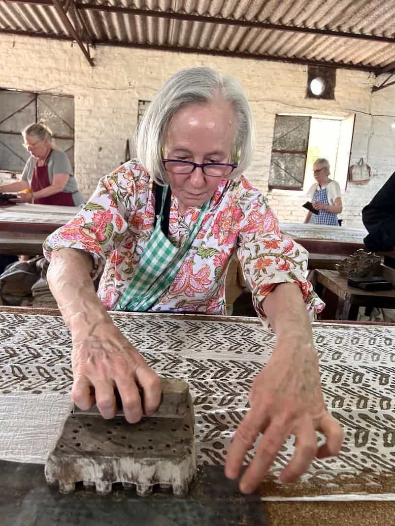 Elderly woman pressing a wooden block onto fabric with intricate patterns in a rustic workshop, participating in a textile resist printing activity.