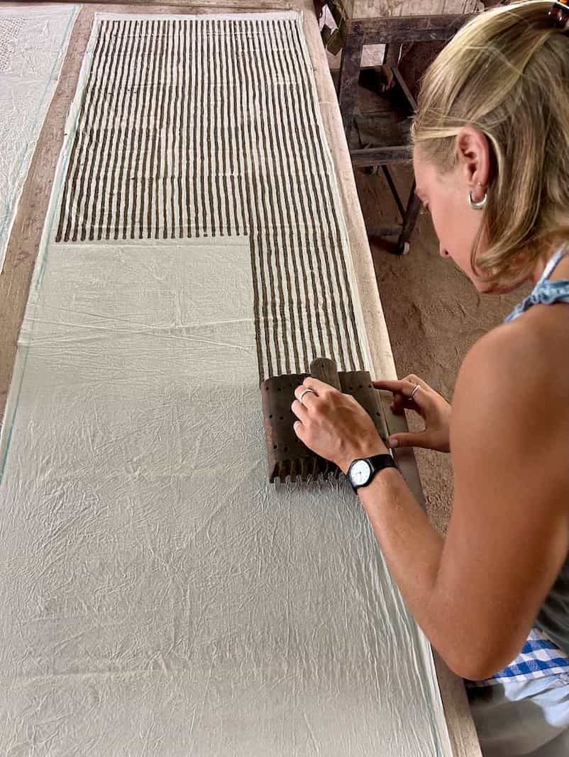 Woman carefully stamping vertical stripe patterns onto a long sheet of fabric using a block in a hands-on resist printing session.