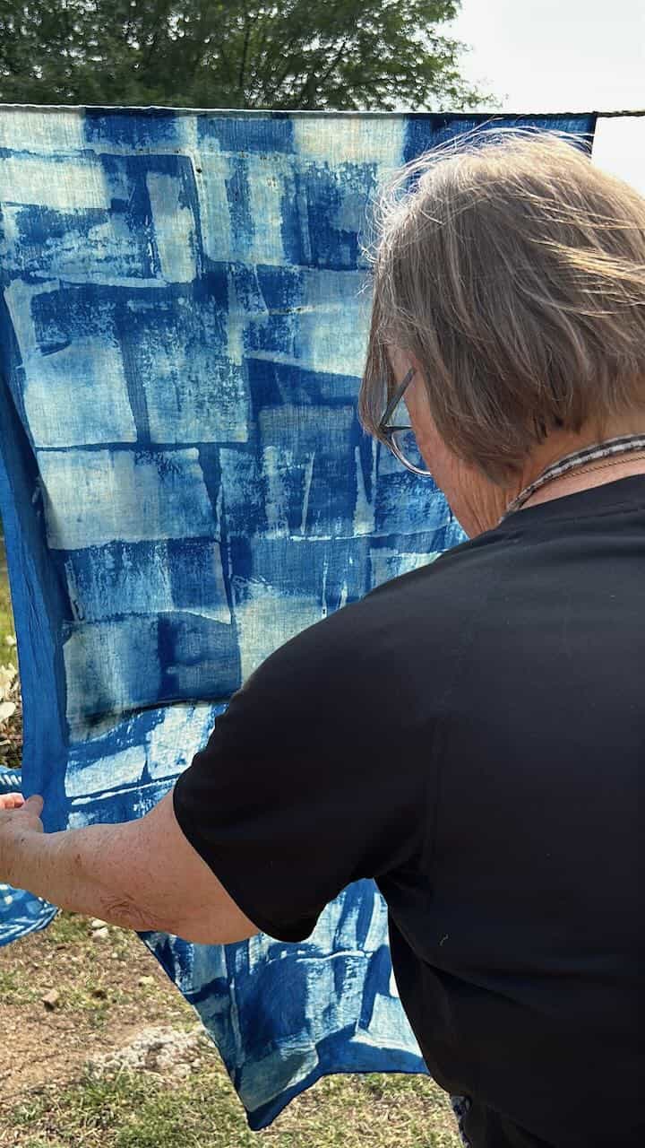 Long pieces of hand-crafted resist-printed fabric hanging outdoors on a line to dry, revealing detailed organic and geometric motifs in earth tones.